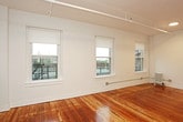 This single-floor unit is characterized by its open layout and warm, polished hardwood flooring that extends throughout the main living areas, reflecting natural light and adding a rich, inviting tone to the space. The expansive main living area is lined with multiple double-hung windows fitted with white roller shades, allowing generous natural light to fill the room while offering views of the surrounding neighborhood. A classic cast-iron radiator provides supplemental heating, and a track lighting system runs along the ceiling, offering flexible illumination. The crisp white walls and clean architectural lines contribute to a bright, airy atmosphere throughout.

The kitchen is efficiently designed, featuring white cabinetry both above and below, stone-look countertops, and a suite of stainless steel appliances including a gas range, dishwasher, refrigerator, and over-the-range microwave. The hardwood flooring continues seamlessly into the kitchen, maintaining visual continuity.

The bathroom is finished with large-format white subway tiles lining the tub surround, paired with chrome fixtures and a clean, minimalist aesthetic. The overall design of the space balances classic architectural character with modern, updated finishes.