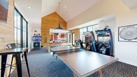 Located in Golden, Colorado, this expansive single-floor game room delivers a comprehensive entertainment experience across a generously sized footprint. The space is divided into two distinct zones seamlessly connected by a hallway. The bowling area commands attention with two full-length regulation lanes flanked by bold crimson accent walls, natural wood paneling, and large picture windows that flood the lanes with natural light and offer views of the surrounding Colorado landscape. Decorative circular porthole-style mirrors add a playful design touch along the walls. The adjacent game lounge features soaring vaulted ceilings with prominent wood-clad gable walls, creating an airy, open atmosphere. A solid wood pool table with a striking red felt surface serves as the centerpiece, complemented by a full-size ping pong table and multiple arcade racing simulators. Patterned blue carpeting grounds the lounge area, while recessed and pendant lighting provide warm, focused illumination. Large floor-to-ceiling windows frame outdoor views, enhancing the bright and energetic ambiance throughout. The overall design blends rustic wood elements with vibrant color accents, making this game room a dynamic and well-equipped recreational destination.
