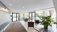 This well-appointed building lobby offers a polished and welcoming first impression, characterized by its clean, contemporary design. Expansive floor-to-ceiling glass walls and large glazed entry doors flood the space with abundant natural light, while providing unobstructed views of the tree-lined exterior and surrounding neighborhood. The open layout is finished with large-format light tile flooring, complemented by a neutral color palette of white and soft grey tones throughout.

A comfortable seating area near the entrance features upholstered lounge chairs and natural wood accent tables, creating an inviting space for residents and guests. Lush potted plants are thoughtfully placed throughout, adding a touch of warmth and life to the modern aesthetic.

Along one wall, a neatly organized bank of individual resident mailboxes is mounted against a contrasting slate-grey accent wall, providing a practical and visually distinct feature. Adjacent to the mailboxes, a staircase with a sleek metal railing leads to the upper floors, while a secured interior door provides access to additional building areas. Recessed ceiling lighting ensures the entire lobby is evenly and warmly illuminated. Overall, the space strikes a balance between functional design and a refined, contemporary ambiance.