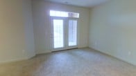 This single-floor space offers a clean, understated layout designed for comfortable everyday living. The main living area is finished with wall-to-wall carpet in a neutral tone, providing a soft and inviting foundation throughout the space. A standout feature is the set of French doors, which serve as the primary source of natural light, flooding the interior with bright, diffused illumination throughout the day. A transom window positioned directly above the French doors further enhances the sense of openness and light. The walls are finished in a soft, warm neutral palette that complements the light carpeting and contributes to the overall airy feel of the space. The room benefits from generous proportions, offering a comfortable sense of scale without feeling overwhelming. Subtle architectural details such as the clean wall lines, visible ceiling vents, and neutral baseboards reflect a practical yet polished interior finish. The French doors provide direct access to an outdoor area, adding a functional connection between the interior and exterior. Overall, the space presents a bright, move-in-ready environment with a calm, neutral aesthetic well-suited for a variety of furnishing styles and uses.