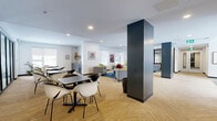 Located at 975 Avenue Saint Charles in Saint-Lambert, Quebec, this generously sized single-floor amenity space offers a thoughtfully designed layout that balances social gathering, relaxation, and recreation. The welcoming entrance lobby features large glass doors, patterned carpet tile flooring, indoor greenery, and stylish accent seating — including a distinctive blue wingback chair and a cognac leather egg chair — setting a polished tone from the moment of arrival. The open-plan main area transitions seamlessly between a casual dining zone with black bistro tables and modern white-and-black chairs, and a comfortable lounge furnished with grey sofas and armchairs accented by bold mustard and red cushions, anchored by a rich blue area rug. Natural light streams through multiple double-hung windows dressed with zebra blinds. Dark charcoal structural columns provide an elegant architectural contrast to the crisp white walls and warm striped carpet flooring throughout. A glass-partitioned multipurpose room with dark-framed sliding panels and hardwood-style flooring offers privacy and flexibility for meetings or events. Adjacent, a games room enclosed in floor-to-ceiling glass features a pool table and bar-height seating. A cozy media room with a textured 3D accent wall, dark ceiling, a wall-mounted TV, and a large sectional sofa completes the space, offering an immersive retreat for residents.