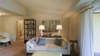 This single-floor residence showcases an open-concept design that integrates the kitchen, dining, and living spaces into one cohesive and functional layout. The kitchen features a light-toned counter that extends as a breakfast bar, providing a natural transition into the dining area. An elegant chandelier with multiple white shades hangs above the dining space, casting warm ambient light and serving as a focal point. A large decorative mirror adorns the wall, enhancing the sense of depth and brightness in the room. The living area is finished with plush carpeting and anchored by a media console housing a flat-screen television. A seating arrangement with a sofa and accent chairs sits nearby, complemented by glass-topped side tables that add a refined touch. Warm beige and taupe tones carry throughout the walls, creating a cohesive and inviting atmosphere. A sliding glass door at the far end of the living area floods the space with natural light and provides access to an outdoor area, adding an airy, open feel to the interior. Overall, the space balances functional design with tasteful décor.
