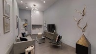 This single-floor space showcases a refined modern aesthetic, characterized by impressively tall ceilings that amplify the sense of openness throughout. The living area is anchored by a contemporary grey sofa, a mirrored coffee table, and a sleek black leather accent chair with chrome legs, all arranged around a flat-screen television positioned atop a minimalist media console. Recessed lighting is complemented by a striking sputnik-style chandelier, adding an artistic focal point overhead. The walls are finished in a cool light grey, contributing to the space's clean, understated palette, while dark hardwood-style flooring grounds the interior with warmth and contrast. A built-in wet bar or kitchenette is seamlessly integrated along one wall, featuring crisp white cabinetry extending to the ceiling for ample storage. A mirrored wall detail in the background adds depth and dimension. Decorative accents, including a natural wood stump side table and curated artwork, lend personality and texture to the otherwise streamlined environment. The overall ambiance is sophisticated, contemporary, and thoughtfully appointed.
