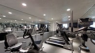 The fitness center is a dedicated single-floor space designed to support a well-rounded cardio and strength training routine. The room is outfitted with a selection of modern exercise equipment, including treadmills, elliptical machines, and stationary bikes, all arranged to maximize usable floor space while maintaining comfortable circulation between machines. A large wall-length mirror spans one side of the room, visually expanding the space and allowing users to monitor their form during workouts. Recessed ceiling lights are evenly distributed throughout, providing consistent, bright illumination across the entire area. The neutral color palette—featuring white walls and light-toned flooring—contributes to a clean, focused atmosphere. Small windows allow natural light to filter in, complementing the overhead lighting. The overall layout is practical and efficient, catering to residents or guests seeking a convenient on-site option for their daily fitness needs without having to leave the building.