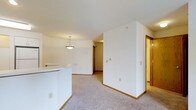 This single-floor space presents an open, functional layout that connects the kitchen and living areas seamlessly. The kitchen features a curved white countertop that extends into the space as a partial divider, complemented by a hanging pendant light with a bowl-shaped shade that adds a focal point overhead. The living area is finished with neutral-toned wall-to-wall carpeting, contributing to a warm and understated ambiance throughout.

The walls are painted a clean, bright white, enhancing the sense of openness and allowing natural and artificial light to reflect evenly across the space. Warm wood-toned baseboards and door trim provide subtle contrast against the white walls and light carpet. A hallway visible in the background leads to additional rooms, with a wood-paneled door adding a traditional touch. A wall-mounted thermostat is conveniently positioned in the main living area. Electrical outlets are thoughtfully placed throughout, indicating practical utility. The overall design is straightforward and neutral, offering a versatile canvas well-suited for a variety of furnishing styles and personal preferences.