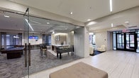 This contemporary amenity space is thoughtfully designed to offer a versatile and engaging environment across a single level. At its centerpiece is a glass-enclosed activity room, defined by full-height frameless glass walls that provide an open, transparent feel while visually connecting the space. Inside, a foosball table serves as a focal point, accompanied by a sleek central counter or bar area and wall-mounted televisions, making it ideal for recreation and social gatherings. Linear LED lighting runs along the ceiling, casting a clean, modern glow throughout the room, while recessed lighting adds additional depth and warmth. The patterned carpet within the glass room contrasts with the light-toned tile flooring of the surrounding areas, creating a clear visual distinction between zones. Adjacent to the glass enclosure, a billiards table and bar-height seating extend the entertainment options further. A separate lounge area features plush cream-toned chairs arranged on a decorative area rug, offering a comfortable retreat for relaxation. Windows allow natural light to filter into the lounge, enhancing the bright and welcoming ambiance. The overall design blends functionality with a polished, upscale aesthetic suited for community or hospitality use.