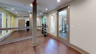 This yoga and fitness studio is a thoughtfully designed single-floor space built to support a variety of movement and wellness practices. The studio features warm hardwood-style flooring throughout, providing a durable yet inviting surface well-suited for yoga, stretching, and light exercise. Recessed lighting is evenly distributed across the ceiling, ensuring consistent, glare-free illumination throughout the practice area.

One of the studio's standout features is the full-length mirrored wall, accompanied by a wall-mounted ballet barre — ideal for alignment-focused practices, barre classes, or dance-inspired workouts. A row of interior windows along one accent wall, painted in a soft, calming yellow-green hue, connects the studio visually to adjacent spaces and allows natural light to filter through, enhancing the open and airy atmosphere.

The studio also includes a dedicated storage area for fitness equipment, with a ball rack visible near the entry, and a yoga mat stored along the wall, reflecting the space's versatility. A glass-paneled door opens to a neighboring fitness area, allowing for seamless transitions between workout zones. Clean white walls, neutral baseboards, and a minimalist aesthetic throughout contribute to a focused, serene environment well-suited to both individual practice and group instruction.