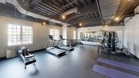Located at 25 Winooski Falls Way in Winooski, Vermont, this fitness center offers a well-appointed, single-floor exercise facility with a generous open layout. The main exercise room is the heart of the space, characterized by an exposed ceiling with dark-painted structural beams, ductwork, and industrial-style cage pendant lighting that lend a modern, loft-like aesthetic. Rubber flooring throughout the workout area provides durability and comfort underfoot.

The exercise room is comprehensively equipped with cardio machines including treadmills, ellipticals, and a stationary bike, as well as free weights, a squat rack with a barbell and weight plates, and a dumbbell rack. A mirrored wall along one side amplifies the sense of space and supports strength training. Yoga mats and resistance accessories are neatly organized along the wall, offering a dedicated stretching and flexibility area. Natural light filters in through multiple large multi-pane windows, brightening the workout environment.

A transitional area near the entrance features warm wood-toned flooring, a built-in water fountain, and a utility storage cart, providing a practical entryway into the facility. The bathroom is clean and functional, equipped with a vanity sink, mirror, soap dispenser, and paper towel dispenser. Together, these spaces form a complete, well-designed fitness facility.