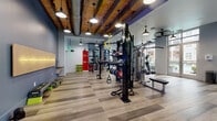 This fitness center at 2120 N Brevard St in Charlotte's NoDa neighborhood offers a thoughtfully designed workout space that blends industrial character with modern functionality. The standout architectural feature is the exposed wood-beam ceiling with visible ductwork, lending a warm, loft-like ambiance complemented by stylish pendant lighting throughout. Wide-plank wood-look flooring runs across the entire floor, providing a clean and durable surface underfoot. Large floor-to-ceiling windows along the perimeter flood the space with natural light and offer open views of the surrounding neighborhood. The fitness floor is comprehensively equipped with a multi-functional cable training rig featuring TRX suspension trainers, battle ropes, and medicine balls, alongside a dedicated free weights area with a full dumbbell rack and flat bench. Cardio equipment includes a row of Precor treadmills with individual screens, ellipticals, and upright bikes. A punching bag station adds a functional training element, while accent walls in deep teal and bold blue add visual energy to the space. Full-length mirrors along one wall enhance the sense of openness. Rustic wooden columns punctuate the space, reinforcing the industrial-chic aesthetic that defines this well-appointed fitness facility.