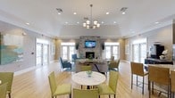 This single-floor clubhouse offers a thoughtfully designed communal space that balances comfort and style. The main lounge area commands attention with its vaulted ceiling and an elegant chandelier that anchors the room, while recessed lighting throughout ensures a bright and inviting atmosphere. Large windows line the perimeter, flooding the interior with natural light and providing open views of the surrounding exterior.

At the heart of the room stands a striking stacked-stone fireplace, topped with a wall-mounted flat-screen television, creating a natural focal point for the space. The seating arrangement is both functional and stylish, featuring a central sofa grouping complemented by accent chairs in teal and warm neutral tones. A round marble-topped table with green upholstered chairs in the foreground provides a casual dining or gathering spot.

The light hardwood flooring runs throughout, lending warmth and cohesion to the overall design. Neutral wall tones paired with clean white trim create a fresh, polished aesthetic. Framed artwork accents the walls, adding subtle personality to the space. With its open layout, generous proportions, and well-curated furnishings, this clubhouse is well-suited for both relaxed gatherings and community events.