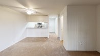 This single-floor studio layout offers a practical and well-organized living space designed for comfort and efficiency. The open living area is finished with plush wall-to-wall carpeting and features a ceiling fan, a wall-mounted mini-split unit for climate control, and a sliding glass door fitted with vertical blinds that invites natural light and provides access to an outdoor area. The kitchen is thoughtfully designed in a galley-style configuration, featuring stainless steel appliances including a refrigerator, range, and over-the-range microwave. Granite-style countertops and white cabinetry provide a clean, functional workspace, while light tile flooring and a skylight-style ceiling panel brighten the space. A pass-through counter connects the kitchen to the living area, enhancing the open feel. The bathroom is well-appointed with a double-sink vanity topped with granite-style countertops, chrome fixtures, a large mirror with overhead lighting, and a towel bar. A separate water closet provides added privacy. Storage is addressed with a closet accessible from the main living area. The overall palette of neutral tones throughout creates a cohesive, move-in-ready environment.