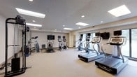 This fitness center offers a clean, well-organized workout environment spread across a single floor. The space is outfitted with a comprehensive selection of exercise equipment, including multiple treadmills, upright stationary bikes, and elliptical or cross-trainer machines, providing ample options for cardiovascular workouts. A dedicated strength and resistance training area features additional machines and free weights, catering to a full range of fitness routines.

The room is finished with neutral-toned walls and light carpeted flooring, creating a comfortable and inviting atmosphere. Overhead, a drop ceiling fitted with large skylight panels and recessed lighting ensures the space is consistently and evenly illuminated throughout the day. Multiple flat-screen televisions are mounted at strategic points around the room, allowing users to stay entertained during their workouts.

Natural light filters in through a large window along one wall, adding to the bright and airy feel of the space. The layout is thoughtfully arranged to maximize open floor space, ensuring easy movement between equipment. Overall, this fitness center provides a well-rounded, functional environment suitable for a variety of workout preferences and fitness levels.