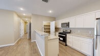 This single-story home offers a well-designed layout accommodating five bedrooms and two full bathrooms. The open-concept kitchen and living area serves as the heart of the home, featuring crisp white shaker-style cabinetry extending to the ceiling, light granite countertops, and a full suite of stainless steel appliances including a gas range, built-in microwave, and a large refrigerator. A central kitchen island with a granite countertop and integrated sink provides additional prep space and casual seating. A dedicated pantry adjacent to the kitchen offers generous built-in shelving for storage. Wood-look tile flooring flows consistently throughout the entire home, offering both durability and a warm aesthetic. The living area is bright and airy, benefiting from multiple windows fitted with white blinds and a ceiling fan. The primary bedroom is a comfortable size and includes a walk-in closet with built-in shelving and hanging rods. The remaining four bedrooms are well-proportioned with neutral warm-toned walls and the same consistent flooring. Both bathrooms are finished with granite-topped white vanities, undermount sinks, and matching wood-look tile floors. The home presents a clean, neutral palette throughout, creating a move-in ready environment suited for a variety of lifestyles.