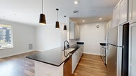 Located at 4540 Helm Dr in Virginia Beach, VA, this single-floor residence offers a thoughtfully designed interior with a clean, contemporary aesthetic throughout. The kitchen serves as a central highlight, featuring dark granite-style countertops, gray shaker cabinetry, a subway tile backsplash, and stainless steel appliances including a refrigerator, range, and microwave. A peninsula with an undermount sink and matte black faucet anchors the space, complemented by three striking black pendant lights overhead. Tall cabinetry extends to the ceiling near the entryway, maximizing storage.

Wood-style plank flooring runs through the main living and kitchen areas, creating a warm, cohesive flow. The living area opens to the outdoors through a glass door flanked by a window, inviting natural light and exterior views. A separate bedroom features floor-to-ceiling corner windows that flood the space with daylight, while additional carpeted rooms offer a quieter, softer feel. Closet space is provided with wire shelving systems.

The bathroom is finished with wood-look flooring, a white vanity with a clean countertop, a large mirror, and matte black hardware and towel bars, maintaining the home's consistent modern palette.
