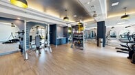 Located in Acworth, Georgia, this contemporary fitness center offers a thoughtfully designed, single-floor exercise facility that accommodates a wide range of workout styles. The space is organized into distinct zones, each tailored to a specific type of training, creating a seamless and functional flow throughout.

The cardio area is bathed in natural light from large floor-to-ceiling windows, offering outdoor views that enhance the workout experience. Multiple treadmills and elliptical machines with integrated screens are positioned to take full advantage of the natural light. Stylish black and gold pendant lights hang from a coffered ceiling with warm LED cove lighting, adding a polished, upscale ambiance.

Adjacent areas feature open-plan functional training zones with hardwood-style flooring, spinning bikes, a tire sled, and TRX suspension systems. A dedicated strength training section houses cable machines, weight benches, and a dumbbell rack, all framed by sleek navy blue accent walls and arched architectural details.

A separate studio room, enclosed by floor-to-ceiling glass-panel doors, provides a quiet space for flexibility and group fitness. It is outfitted with exercise balls, medicine balls, resistance bands, and a large wall mirror paired with a mounted display screen. Neutral textured wall panels and recessed LED cove lighting complete the refined aesthetic throughout the entire facility.