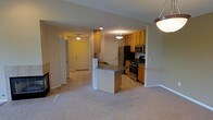 This single-floor residence showcases an open-concept design that connects the living area and kitchen, creating a cohesive and functional layout. The kitchen is well-appointed with stainless steel appliances, including a gas range, microwave, and refrigerator, all complemented by warm honey-toned cabinetry that provides ample storage. A peninsula with a dark granite-style countertop extends into the space, offering additional prep space and casual seating opportunities.

The flooring transitions from light ceramic tile in the kitchen and entryway to plush carpeting in the living area, adding warmth and comfort underfoot. Recessed lighting and pendant fixtures throughout ensure consistent, well-distributed illumination across both spaces. The neutral wall tones enhance the bright, open feel of the interior.

A clearly defined entryway with a white front door provides a welcoming arrival point, while an adjacent hallway leads to additional rooms beyond. A fireplace is partially visible along the living room wall, adding a notable architectural feature. The overall ambiance is warm and practical, with a layout that balances everyday functionality with comfortable living.