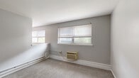 Located at 8223 Roosevelt Blvd in Philadelphia, PA, this single-floor unit offers four bedrooms and one bathroom within a compact, efficiently arranged floor plan. The layout, as seen in the dollhouse view, organizes the rooms in a straightforward linear configuration, maximizing the use of available space.

Each bedroom features wall-to-wall carpeting in a neutral beige tone and is painted in a consistent light gray palette, creating a clean and cohesive look throughout. The bedrooms are well-lit thanks to multiple windows fitted with white horizontal blinds, allowing natural light to filter in while maintaining privacy. Baseboard heating runs along the walls, and select rooms are also equipped with window air conditioning units for year-round climate control. Several bedrooms include built-in closets with hanging rods, providing practical storage.

The spacious primary bedroom stands out for its generous proportions, dual windows, and open feel, with a sightline into the adjacent kitchen area. The kitchen features warm wood-toned cabinetry, visible from the primary bedroom doorway.

The bathroom is functional and neatly appointed, with a white vanity, mirrored medicine cabinet, toilet, and tile-like wainscoting along the lower walls, complemented by durable flooring. Overall, the unit presents a clean, move-in ready interior well-suited for practical everyday living.