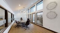 This contemporary conference room is thoughtfully designed to support productive meetings in a polished, professional environment. The space is defined by its striking vaulted ceilings, which lend an impressive sense of openness and volume. A row of large, dark-framed windows lines one wall, flooding the interior with natural light and offering views to an adjacent outdoor terrace, accessible via a glass-paneled door — providing an optional extension of the meeting space.

At the center of the room sits a substantial rectangular conference table with built-in power access points, surrounded by a mix of executive and accent chairs in complementary tones. Overhead, a sculptural multi-bulb chandelier serves as a focal point, adding warmth and a modern aesthetic to the bright white walls. Recessed lighting supplements the natural and ambient light throughout.

Textured, striped carpeting grounds the space and adds a layer of acoustic comfort. Wall-mounted geometric art pieces and a branded interior window panel contribute to the room's distinct, curated character. A secondary alcove area adjacent to the main conference space features additional wall art and a mid-century-style display cabinet, adding depth and a refined finishing touch to this well-appointed professional space.