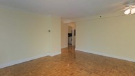 This single-floor space showcases a clean, understated design throughout. The main living area is an open, rectangular room finished with warm parquet wood flooring in a classic block pattern, adding visual texture and character underfoot. Crisp white walls and white crown molding frame the space, contributing to a bright and airy atmosphere. The neutral color palette creates a versatile backdrop suited to a variety of furnishing styles.

A wide open doorway connects the living area to the kitchen, which is visible in the background. The kitchen features white cabinetry, a white range, and a full-sized refrigerator, offering functional cooking amenities. The layout allows for easy flow between the two areas.

White baseboard trim runs consistently throughout, complementing the overall clean aesthetic. The space receives adequate interior lighting and maintains a straightforward, practical layout. Overall, the unit presents a simple yet classic interior defined by its warm hardwood parquet floors, neutral tones, and efficient use of connected living and kitchen spaces.