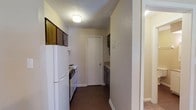 Located at 2436 Beach Blvd in Biloxi, Mississippi, this single-floor unit offers a straightforward, functional layout. The living area serves as the primary gathering space, finished with wall-to-wall carpet, a ceiling fan with light fixture, a wall-mounted HVAC unit, and a window that brings in natural light. A panel entry door provides direct access to the exterior.

The kitchen is positioned adjacent to the living area and is equipped with essential appliances, including a white refrigerator, an electric coil range, and a dishwasher. Dark wood cabinetry overhead provides storage, while a double-basin stainless steel sink and laminate countertops complete the workspace. Dark vinyl flooring runs throughout the kitchen, contrasting with the carpeted living space.

The bathroom features wood-look vinyl flooring and includes a full tub-shower combination with a molded surround, a toilet, and a towel bar. A bifold door opens to an adjacent closet, offering practical storage. Textured ceilings are consistent throughout the unit, contributing to its utilitarian character. The overall layout is compact and efficiently organized, making practical use of the available space.