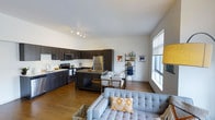 This single-floor space showcases a thoughtfully designed open-concept layout that seamlessly integrates the kitchen, dining, and living areas into one cohesive environment. The kitchen is outfitted with dark espresso cabinetry, stainless steel appliances including a refrigerator, stove, and microwave, and a dark countertop island that doubles as a casual dining or prep space. A dedicated dining area sits adjacent to the kitchen, providing a defined yet connected space for meals. The living area features warm hardwood-style flooring that extends throughout, anchoring a comfortable seating arrangement. Large windows along one wall allow abundant natural light to fill the space, enhancing the open and airy atmosphere. White walls provide a clean, neutral backdrop that contrasts effectively with the darker kitchen finishes and the warm tones of the flooring. Track lighting on the ceiling offers additional illumination in the kitchen and living zones. A lean shelving unit in the background serves as a functional display area, adding a touch of personality to the space. Overall, the layout is efficient and modern.