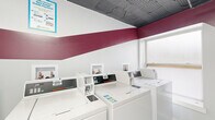 This shared laundry room is a well-organized, single-level facility designed to accommodate communal laundry needs efficiently. The space is outfitted with multiple Speed Queen commercial top-load washing machines arranged in a row along one wall, each with its own dedicated hookup niche built into the wall above. Complementing the washers, stacked commercial dryer units line an adjacent wall, maximizing the use of vertical space and providing ample drying capacity.

The room features light tile flooring, making it easy to maintain, while the walls are painted white with a bold burgundy stripe that wraps around the room, giving the space a clean and distinctive look. An exposed ceiling with visible ductwork and overhead flush-mount lighting provides functional illumination throughout. A wall-mounted circulation fan supports ventilation and air circulation.

A folding table offers a convenient surface for sorting and folding laundry, and a wire laundry cart is available for transporting loads. Informational signage is posted throughout, including notices for the PayRange mobile payment system, which enables cashless operation of the machines. A fire extinguisher is mounted near the entrance, ensuring safety compliance. A frosted window allows natural light to filter into the space while maintaining privacy.