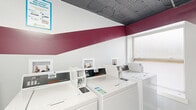 This shared laundry room is a well-organized, single-level facility designed to accommodate communal laundry needs efficiently. The space is outfitted with multiple Speed Queen commercial top-load washing machines arranged in a row along one wall, each with its own dedicated hookup niche built into the wall above. Complementing the washers, stacked commercial dryer units line an adjacent wall, maximizing the use of vertical space and providing ample drying capacity.

The room features light tile flooring, making it easy to maintain, while the walls are painted white with a bold burgundy stripe that wraps around the room, giving the space a clean and distinctive look. An exposed ceiling with visible ductwork and overhead flush-mount lighting provides functional illumination throughout. A wall-mounted circulation fan supports ventilation and air circulation.

A folding table offers a convenient surface for sorting and folding laundry, and a wire laundry cart is available for transporting loads. Informational signage is posted throughout, including notices for the PayRange mobile payment system, which enables cashless operation of the machines. A fire extinguisher is mounted near the entrance, ensuring safety compliance. A frosted window allows natural light to filter into the space while maintaining privacy.