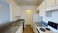 This single-floor space is centered around a well-organized galley-style kitchen designed for efficient use of space. The kitchen features white cabinetry lining both walls, providing ample upper and lower storage. Gray speckled laminate countertops run along each side, offering generous prep space. On one side, a double-basin stainless steel sink is paired with a dishwasher, while the opposite side hosts a white electric coil range and additional counter space. A full-size white refrigerator anchors one end of the kitchen, positioned beside a doorway that leads to a dedicated laundry area housing a stacked washer and dryer unit. The kitchen floor is finished with warm wood-tone laminate planking that adds a touch of warmth to the clean, neutral palette. Overhead, a ceiling vent ensures proper ventilation throughout the space. The overall design is clean and practical, with a bright, neutral color scheme of white cabinetry against light-toned walls that creates an open and airy feel. The layout maximizes functionality, making efficient use of the available footprint while keeping all essential appliances and storage within easy reach.