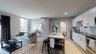 This single-floor residence at 5040 Flats offers a thoughtfully designed open-concept layout finished in a consistent cool-gray palette with light hardwood-style flooring flowing throughout the main living areas. The welcoming entryway features a natural wood console table, a large round mirror, and curated wall art, setting a modern yet personable tone from the moment you step inside.

The open living and dining area is generously proportioned, bathed in natural light from oversized windows and a glass balcony door. The living room comfortably accommodates a full seating arrangement and media setup, with a wall-mounted mini-split system providing climate control. A freestanding kitchen island with a white quartz top bridges the living space and kitchen, offering additional seating and prep space.

The kitchen is well-equipped with white shaker cabinetry, dark countertops, stainless steel appliances including a dishwasher, range, microwave, and refrigerator, and ample storage throughout.

The bedroom is a calm retreat featuring corner windows that invite natural light, plush carpeting, a wall-mounted mini-split unit, a double closet, and generous space for furnishings. A full bathroom is accessible nearby, completing this efficiently planned and contemporary residence.