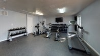 This fitness center is a purpose-built, single-floor exercise facility designed to accommodate a wide range of workout routines. The space features durable rubber flooring throughout, providing both safety and comfort underfoot during workouts. Fluorescent overhead lighting ensures the entire area is well-lit, while light gray walls contribute to a clean, open atmosphere.

The cardio zone is equipped with a treadmill, elliptical trainer, stationary upright bike, and a spin bike, offering diverse options for cardiovascular training. A wall-mounted flat-screen television faces the cardio equipment, enhancing the workout experience. For strength training, a multi-tiered dumbbell rack holds an extensive selection of free weights, and a cable machine provides additional resistance training options. A adjustable workout bench is also available for versatile strength exercises. Kettlebells are stored on a dedicated rack along one wall.

A large wall mirror spans one side of the space, allowing users to monitor their form during exercise. Two water fountains are conveniently mounted along the mirrored wall, and restroom access is available directly from the fitness room. Double glass doors on one end of the space allow natural light to filter in and provide views of the surrounding greenery, creating a welcoming entry point to the facility.
