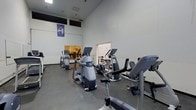 This fitness center offers a comprehensive workout environment designed to accommodate a range of exercise needs. The space is characterized by its impressively high ceilings, which lend an open and airy atmosphere, reducing any sense of confinement commonly found in gym settings. The main workout floor is finished with durable, easy-to-maintain flooring suitable for heavy equipment and high-traffic use. A prominent collection of modern cardio machines, including elliptical trainers and upright cycles, is arranged to allow comfortable spacing between users. A secondary area, visible through a connecting doorway, houses additional equipment including cable machines and strength training stations, expanding the overall capacity of the facility. The neutral white walls keep the environment bright and focused, while a branded pennant adds a touch of community identity to the space. Overhead lighting ensures consistent illumination throughout. The layout is thoughtfully organized to create distinct zones for different types of exercise, making navigation intuitive for users of all fitness levels. Overall, the fitness center provides a clean, functional, and well-maintained setting suited for daily training use.