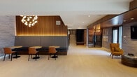 This thoughtfully designed amenity space blends contemporary style with functional comfort across a single, well-appointed floor. The open social lounge serves as the heart of the space, anchored by a striking linear fireplace set into a white brick feature wall, complemented by a warm wood-paneled ceiling, built-in shelving, and plush sectional seating arranged around a curated area rug.

Adjacent to the lounge, a fully equipped kitchen features rich dark wood cabinetry, stainless steel appliances, a tiled backsplash, and an extended bar-height island illuminated by a row of gold globe pendant lights — ideal for casual gatherings. A separate dining area with a bold patterned ceiling detail and floor-to-ceiling black-framed glass doors opens directly to an outdoor terrace and pool area, flooding the interior with natural light and outdoor views.

The main corridor connects additional seating vignettes, including a cozy TV nook with mustard accent chairs, warm wood ceiling, and textured stone wall cladding. A dedicated gaming room with carpeted flooring and recessed lighting rounds out the recreational offerings. Light hardwood floors, a sculptural chandelier, and built-in banquette seating in the entry area tie the entire space together with a cohesive, upscale aesthetic.