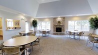 This single-story clubhouse is designed to foster community gathering in a bright, open environment. The main gathering area is defined by a dramatic vaulted ceiling that rises prominently above the space, creating an airy and expansive atmosphere. Neutral tones throughout the walls and flooring lend the room a clean, understated elegance.

A floor-to-ceiling stone fireplace serves as the room's central focal point, adding warmth and a classic architectural character. Its natural stacked-stone construction is complemented by a decorative accent piece mounted above the mantle. Large multi-pane windows line the walls on either side of the fireplace, flooding the interior with generous natural light and providing views of the surrounding greenery.

The space is furnished with multiple round tables and lightweight chairs arranged throughout, accommodating a variety of group sizes and event configurations. A decorative indoor tree adds a touch of organic warmth to the corner of the room. Recessed lighting supplements the natural light, ensuring the space remains well-lit at all hours. Neutral carpeted flooring ties the room together, creating a comfortable and inviting environment suitable for a wide range of community activities.