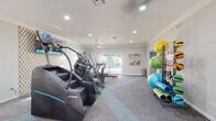 Located at 4031 Vista Verde Dr in New Port Richey, FL, this well-appointed fitness center is a single-floor facility designed to support a wide range of workout routines. The space is finished with crown molding, recessed lighting, and ceiling fans that contribute to a clean, modern aesthetic throughout.

The main exercise area is generously sized and outfitted with True brand stair climbers, elliptical machines, treadmills, and a stationary bike, all arranged to allow comfortable use without crowding. Patterned accent wallpaper adds a stylish touch alongside the neutral gray carpeted flooring, which features teal geometric detailing for visual interest.

A dedicated free-weight zone includes a full rack of dumbbells and a weight bench, accompanied by a decorative gold-toned open shelving unit along the wall. Natural light filters in through blinds-covered windows, brightening the space during daytime hours.

A well-stocked equipment storage rack holds medicine balls, resistance bands, stability balls in multiple sizes, and rolled exercise mats, making group or solo functional training accessible. Sliding glass doors open directly to an outdoor pool and patio area, creating a seamless indoor-outdoor connection. A separate, smaller adjacent room — visible through a glass-panel door — appears to serve as a private office or consultation space. Full-length mirrors and a large wall-mounted television round out the facility's practical amenities.