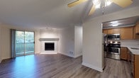 This single-floor residence offers a thoughtfully designed layout with a blend of comfort and functionality throughout. The living room serves as the central gathering space, anchored by a classic tile-surround fireplace with a clean white mantel, track lighting, and a ceiling fan. Large sliding glass doors open to a private balcony, flooding the room with natural light and providing views of the surrounding greenery. The flooring throughout the main living areas features durable hardwood-style planks in a warm grey-brown tone, lending a modern, cohesive aesthetic.

The kitchen is efficiently arranged with warm honey-toned wood cabinetry, light stone countertops, and a full suite of stainless steel appliances including a refrigerator, range, and over-the-range microwave. The adjacent entry area features a ceiling fan and a paneled entry door. The bedrooms offer plush carpeting, large windows with vertical blinds for natural light and privacy, and baseboard heating units. Crown molding details are present in the main living areas, adding a polished finishing touch. The bathroom is cleanly finished with a tub-shower combo, white tile surround, and a warm wood vanity with a light stone countertop.