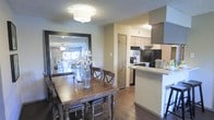 This single-floor space showcases an open-concept layout that seamlessly connects the dining area and kitchen. The dining space is anchored by a rustic wood dining table paired with dark crossback chairs, complemented by a large decorative mirror that reflects light and enhances the sense of depth in the room. A flush-mount ceiling light fixture illuminates the area, while a textured ceiling adds a classic touch overhead. Dark hardwood flooring runs throughout, providing a warm, cohesive foundation.

The adjacent kitchen features white upper cabinetry, a mosaic tile backsplash, and a raised breakfast bar that offers additional counter space and casual seating. Black appliances, including a refrigerator and range, provide a striking contrast against the lighter cabinetry and countertops. A range hood adds both function and structure above the cooking area. A tall pantry-style cabinet offers practical built-in storage. The kitchen's layout is efficient and accessible, with the peninsula bar serving as a natural divider between the culinary and dining zones. Overall, the space presents a functional and inviting atmosphere with a blend of traditional and contemporary design elements.
