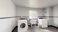 This shared laundry facility is a clean, well-organized single-floor space designed for practical, everyday use. The room is finished with crisp white walls accented by a navy blue horizontal stripe running along the lower portion, complemented by matching navy baseboards and door trim that give the space a cohesive, institutional look. The flooring throughout is a durable speckled tile, easy to maintain and suited to heavy use.

The laundry area is equipped with commercial-grade front-loading and top-loading washing machines alongside matching dryers, providing ample capacity. A freestanding utility sink is centrally positioned near the window, offering convenient access for hand-washing or pre-treatment. A wall-mounted folding table provides a generous surface for sorting and folding laundry, making the space both efficient and user-friendly.

Natural light fills the room through a large window overlooking mature trees, creating a pleasant and airy atmosphere. A blue steel door with a small window panel provides secure entry and exit. Informational postings are displayed on the walls, and a waste bin is conveniently located near the machines. The layout is open and spacious, allowing multiple users to move comfortably throughout the room simultaneously.