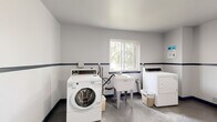 This shared laundry facility is a clean, well-organized single-floor space designed for practical, everyday use. The room is finished with crisp white walls accented by a navy blue horizontal stripe running along the lower portion, complemented by matching navy baseboards and door trim that give the space a cohesive, institutional look. The flooring throughout is a durable speckled tile, easy to maintain and suited to heavy use.

The laundry area is equipped with commercial-grade front-loading and top-loading washing machines alongside matching dryers, providing ample capacity. A freestanding utility sink is centrally positioned near the window, offering convenient access for hand-washing or pre-treatment. A wall-mounted folding table provides a generous surface for sorting and folding laundry, making the space both efficient and user-friendly.

Natural light fills the room through a large window overlooking mature trees, creating a pleasant and airy atmosphere. A blue steel door with a small window panel provides secure entry and exit. Informational postings are displayed on the walls, and a waste bin is conveniently located near the machines. The layout is open and spacious, allowing multiple users to move comfortably throughout the room simultaneously.
