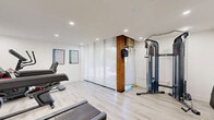 Located at 552 Birchmount Rd in Toronto, Ontario, this dedicated exercise room offers a focused and functional fitness environment within a compact, efficiently utilized single-floor layout. The space is finished with a clean, modern aesthetic, featuring crisp white walls, recessed ceiling lighting, and light-toned laminate flooring that together create a bright and energizing atmosphere.

The room is equipped with a range of cardio and strength training equipment, including a treadmill, a stationary bike, and an elliptical machine, all positioned to allow comfortable use. A wall-mounted flat-screen television faces the cardio equipment, providing entertainment during workouts. On the opposite end of the room, a dual-cable functional trainer offers versatile strength and resistance training options. A foldable exercise mat is also available for floor-based workouts.

A dedicated exercise chart poster is displayed on the wall, serving as a practical reference guide for users. An informational notice is posted near the entrance, indicating shared or regulated use of the space. The entry door features a door closer mechanism, reinforcing the room's purpose as a controlled-access fitness facility. An exposed brick column adds a subtle industrial character to an otherwise clean, contemporary interior, giving the space a unique visual contrast.