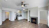 This single-floor space showcases an open-concept design that seamlessly connects the living area and kitchen, creating a spacious and functional layout. The living area is anchored by a gas fireplace set within a clean, white-trimmed surround, flanked by a built-in shelving niche that adds both storage and architectural interest. A ceiling fan with an integrated light fixture is centrally mounted, enhancing air circulation and providing ambient lighting throughout the space. Crown molding runs along the ceiling perimeter, lending a refined, polished finish to the neutral gray walls.

Wood-style plank flooring extends throughout, offering a warm, contemporary aesthetic that ties the open floor plan together cohesively. The kitchen features white cabinetry, stainless steel appliances including a full-size refrigerator, a peninsula with a sink and dark countertops, and subway tile backsplash. Recessed and flush-mount lighting illuminate the kitchen area effectively. The overall palette — soft grays, crisp white trim, and warm wood tones — creates a modern yet inviting ambiance, well-suited for both everyday living and entertaining.