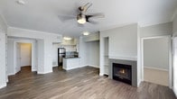 This single-floor space showcases an open-concept design that seamlessly connects the living area and kitchen, creating a spacious and functional layout. The living area is anchored by a gas fireplace set within a clean, white-trimmed surround, flanked by a built-in shelving niche that adds both storage and architectural interest. A ceiling fan with an integrated light fixture is centrally mounted, enhancing air circulation and providing ambient lighting throughout the space. Crown molding runs along the ceiling perimeter, lending a refined, polished finish to the neutral gray walls.

Wood-style plank flooring extends throughout, offering a warm, contemporary aesthetic that ties the open floor plan together cohesively. The kitchen features white cabinetry, stainless steel appliances including a full-size refrigerator, a peninsula with a sink and dark countertops, and subway tile backsplash. Recessed and flush-mount lighting illuminate the kitchen area effectively. The overall palette — soft grays, crisp white trim, and warm wood tones — creates a modern yet inviting ambiance, well-suited for both everyday living and entertaining.