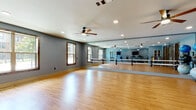 This single-level fitness facility is divided into two distinct zones designed to support a variety of workout styles. The studio space is a standout feature, boasting a wide-plank hardwood floor that reflects natural light streaming in from multiple windows. Floor-to-ceiling mirrors line an entire wall, creating a visually expansive environment ideal for dance, yoga, barre, or group fitness classes. Horizontal ballet barres are mounted in front of the mirrors, and exercise balls and resistance equipment are stored neatly along the perimeter. Two ceiling fans circulate air throughout the open, unobstructed floor plan, while recessed lighting supplements natural illumination.

Adjacent to the studio, the cardio and strength training area features plush patterned carpet and is equipped with treadmills, stationary bikes, rowing machines, a cable weight machine, a full dumbbell rack with a weight bench, and a wall-mounted flat-screen television. A ceiling fan and recessed lighting maintain a comfortable atmosphere. Rustic dark-wood double doors connect the two areas, adding a warm design contrast to the cool blue-grey walls that unify the overall aesthetic of the facility.