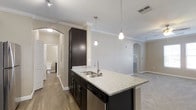 This single-story retreat offers a thoughtfully designed layout that balances functionality with modern aesthetics. The kitchen serves as a central highlight, featuring a peninsula with granite countertops, a stainless steel sink, and rich dark cabinetry that extends to the ceiling for ample storage. Two pendant lights hang above the peninsula, providing focused task lighting while adding a contemporary decorative touch. The kitchen flows seamlessly into the living area, which is framed by elegant crown molding and neutral-toned walls that create a bright, welcoming atmosphere. Arched doorways add an architectural detail that lends a refined character to the space. Light wood-style flooring runs throughout, complementing the overall neutral color palette. The bathroom, visible through a hallway, is clean and functional. The open-concept design between the kitchen and living areas enhances the sense of spaciousness, making the layout feel larger than its footprint. The combination of granite surfaces, dark cabinetry, and soft wall tones gives this retreat a polished, move-in-ready appeal suitable for comfortable everyday living.