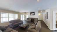 This single-floor space is designed as a comfortable and inviting communal gathering area. The main lounge is generously sized, anchored by a striking stone fireplace with a classic white shiplap surround that serves as the room's focal point. A wall-mounted flat-screen television is positioned above the fireplace, making the space ideal for both relaxation and entertainment. The seating arrangement includes multiple plush sofas and upholstered accent chairs with decorative patterned fabric, all centered around a tufted leather ottoman that doubles as a coffee table. The room features dark carpeting that adds warmth and contrast against the light-painted walls, creating a cozy yet refined ambiance. Recessed ceiling lighting, combined with wall sconces, provides layered illumination throughout the space. Large windows dressed with horizontal blinds line the walls, allowing ample natural light to flow in and offering views of the exterior. A wide doorway connects seamlessly to an adjacent hallway or room, suggesting additional functional areas nearby. Overall, the space strikes a balance between casual comfort and a well-appointed communal environment suited for socializing or unwinding.