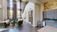 The Heron Downtown Clubhouse is a thoughtfully designed community gathering space that impresses from the moment you enter. The lounge area anchors the room with soaring double-height ceilings and floor-to-ceiling windows dressed in flowing linen curtains, flooding the space with natural light and offering open views of the surrounding neighborhood. A mix of plush seating — including a sleek white sofa accented with vibrant orange throw pillows, teal accent chairs, and a rich green velvet armchair — is arranged atop a bold, colorful area rug, creating an inviting and eclectic atmosphere.

Dark hardwood-style flooring runs throughout, providing a warm contrast to the neutral wall tones. A compact dining nook with a round table and wood-framed chairs sits beneath a wall-mounted flat-screen TV, ideal for casual gatherings. The fully equipped kitchen features sleek gray cabinetry, a green tile backsplash, stainless steel appliances, and a central island with bar seating, topped by a striking geometric pendant light. A large-scale abstract mural in warm gold and amber tones spans the wall above the cabinetry, serving as a bold artistic centerpiece. Together, these elements create a vibrant, welcoming clubhouse with a strong sense of contemporary style.
