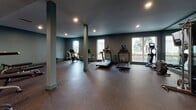 Located at 437 25th St in Oakland, CA, this fitness center is a thoughtfully designed, single-floor facility built to accommodate a wide range of workout needs. The space is defined by its open layout, dark tonal color palette, and rubber-floored surfaces that provide both durability and comfort underfoot.

The cardio zone is generously appointed with multiple treadmills, stationary bikes, and an elliptical machine, all positioned near large windows that allow natural light to filter in and offer views of the surrounding outdoor environment. A dedicated cycling area features Peloton-style bikes on padded mats, along with a rowing machine, creating a focused space for high-intensity sessions.

The strength training section is equally comprehensive, housing a full rack of dumbbells, kettlebells, weight benches, medicine balls, resistance rollers, and a cable machine. Floor-to-ceiling mirrors line the walls in this area, amplifying the sense of space and allowing users to monitor their form. A wall-mounted flat-screen TV provides entertainment during workouts.

Recessed lighting throughout maintains a sleek, modern ambiance, while structural columns punctuate the open floor plan without impeding movement. A water station with dual drinking fountains is conveniently located near the entrance, ensuring easy access to hydration. The overall design balances functionality with a clean, professional aesthetic.