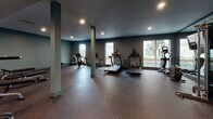 Located at 437 25th St in Oakland, CA, this fitness center is a thoughtfully designed, single-floor facility built to accommodate a wide range of workout needs. The space is defined by its open layout, dark tonal color palette, and rubber-floored surfaces that provide both durability and comfort underfoot.

The cardio zone is generously appointed with multiple treadmills, stationary bikes, and an elliptical machine, all positioned near large windows that allow natural light to filter in and offer views of the surrounding outdoor environment. A dedicated cycling area features Peloton-style bikes on padded mats, along with a rowing machine, creating a focused space for high-intensity sessions.

The strength training section is equally comprehensive, housing a full rack of dumbbells, kettlebells, weight benches, medicine balls, resistance rollers, and a cable machine. Floor-to-ceiling mirrors line the walls in this area, amplifying the sense of space and allowing users to monitor their form. A wall-mounted flat-screen TV provides entertainment during workouts.

Recessed lighting throughout maintains a sleek, modern ambiance, while structural columns punctuate the open floor plan without impeding movement. A water station with dual drinking fountains is conveniently located near the entrance, ensuring easy access to hydration. The overall design balances functionality with a clean, professional aesthetic.