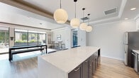 This thoughtfully designed single-floor amenity space blends contemporary style with functional, social-oriented living. The open-concept kitchen serves as a central hub, showcasing an expansive quartz waterfall island, dark cabinetry contrasted by crisp white upper cabinets, herringbone tile backsplash, stainless steel appliances, and a striking cluster of globe pendant lights overhead. Light wood-tone flooring flows consistently throughout, unifying each distinct area.

The lounge area features tray ceilings with crown molding, recessed lighting, and a dramatic focal-point fireplace framed in herringbone tile with a wall-mounted television above. Floor-to-ceiling sliding glass doors flood the space with natural light while providing seamless access to a landscaped outdoor terrace, complete with pergola structures and lush planters.

Adjacent to the lounge, a dedicated billiards room accommodates a full-sized pool table alongside bar-height tables and stools, all bathed in abundant natural light from oversized windows overlooking the terrace. Throughout, the design maintains a cohesive neutral palette of soft whites and grays, delivering a polished, inviting atmosphere well-suited for both relaxation and social gatherings.