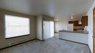 This single-floor space presents an open-concept layout that seamlessly connects the living area and kitchen. The living space is finished with neutral beige carpet and smooth, light-colored walls, creating a clean and understated aesthetic. A large window fitted with horizontal blinds allows natural light to filter in, while a sliding glass door with vertical blinds provides access to an outdoor area, further brightening the interior. A baseboard heater runs along the living room wall, offering a practical heating solution.

The kitchen is positioned along one side of the space and features a peninsula-style counter with a dark laminate surface, offering additional workspace and a natural division between the cooking and living areas. Dark wood cabinetry is mounted above, and a white refrigerator is visible alongside a sink integrated into the counter. Recessed and flush-mounted ceiling fixtures provide consistent overhead lighting throughout. The overall ambiance is simple, functional, and well-lit, with a cohesive neutral palette carried throughout the space.