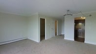 This single-floor space presents a straightforward and functional layout, characterized by neutral, light beige walls and wall-to-wall carpeting that lends a soft, understated warmth throughout. The main room is generously proportioned, providing an open and airy feel despite its simplicity. A classic pendant light fixture hangs from the smooth white ceiling, adding a subtle decorative element to the otherwise understated space.

Two white panel doors are visible along the back wall, one leading to what appears to be a walk-in closet or storage area and the other opening to a smaller closet with visible wood-toned shelving or cabinetry inside. These built-in storage features add practical value to the space. A baseboard heater runs along the lower portion of the back wall, indicating an electric heating system suited to colder climates.

The clean white trim and door casings contrast neatly with the beige walls, contributing to a tidy, classic aesthetic. The space feels move-in ready, offering a blank canvas that accommodates a variety of furnishing styles. Overall, the layout is efficient and well-maintained, with storage and comfort as key functional highlights.
